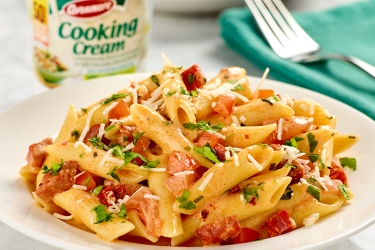 An image of a bowl of penne pasta, tomatoes and chorizo with a bowl of cooking cream in the background